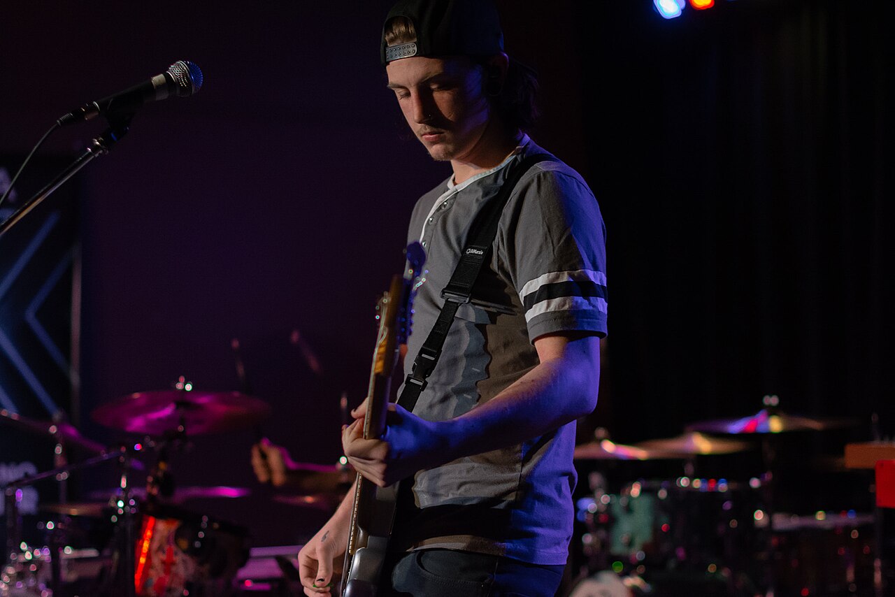 Independent musician performing live on stage with a guitar and microphone, bathed in stage lights, engaging a small but attentive crowd.
